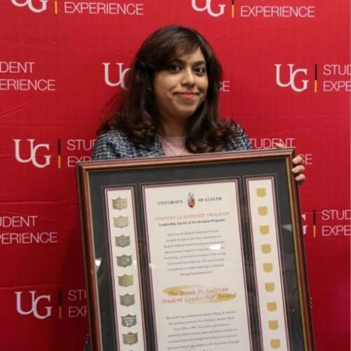 Shaiza holding her large framed award while standing in front of a red U of G Student Experience backdrop and smiling.