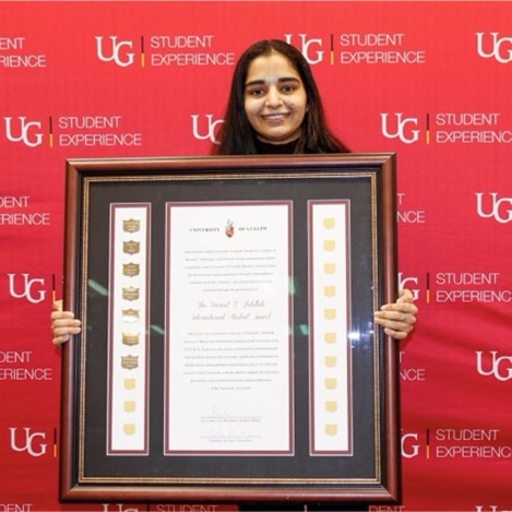 Simran holding their large framed award while standing in front of a red U of G Student Experience backdrop and smiling.