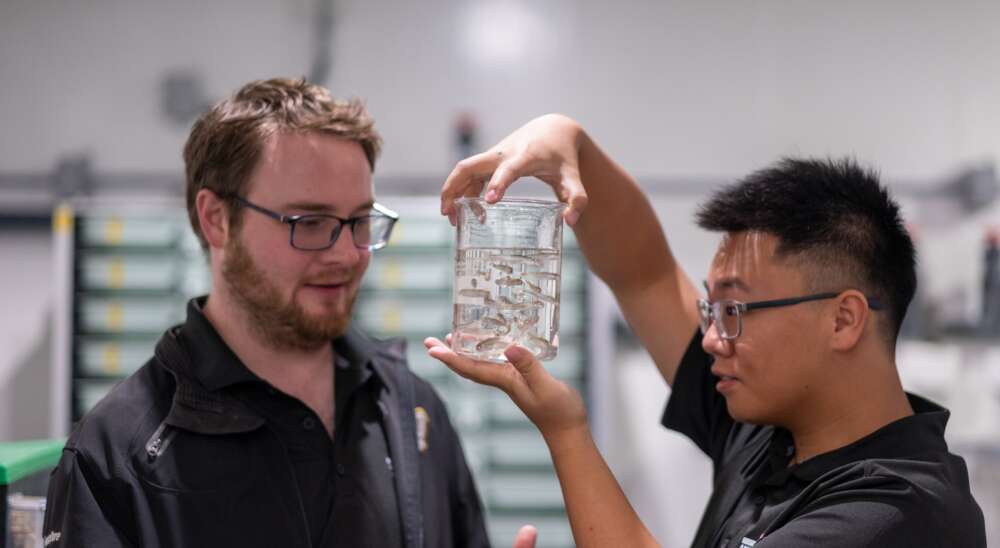 Two researchers wearing black golf shirts stand facing one another while one holds a glass container with water and silver fish.