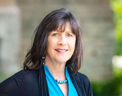 A person with shoulder-length black hair stands on an angle in front of a stone wall looking into the camera wearing a turquoise shirt and necklace with a black sweater over it. 