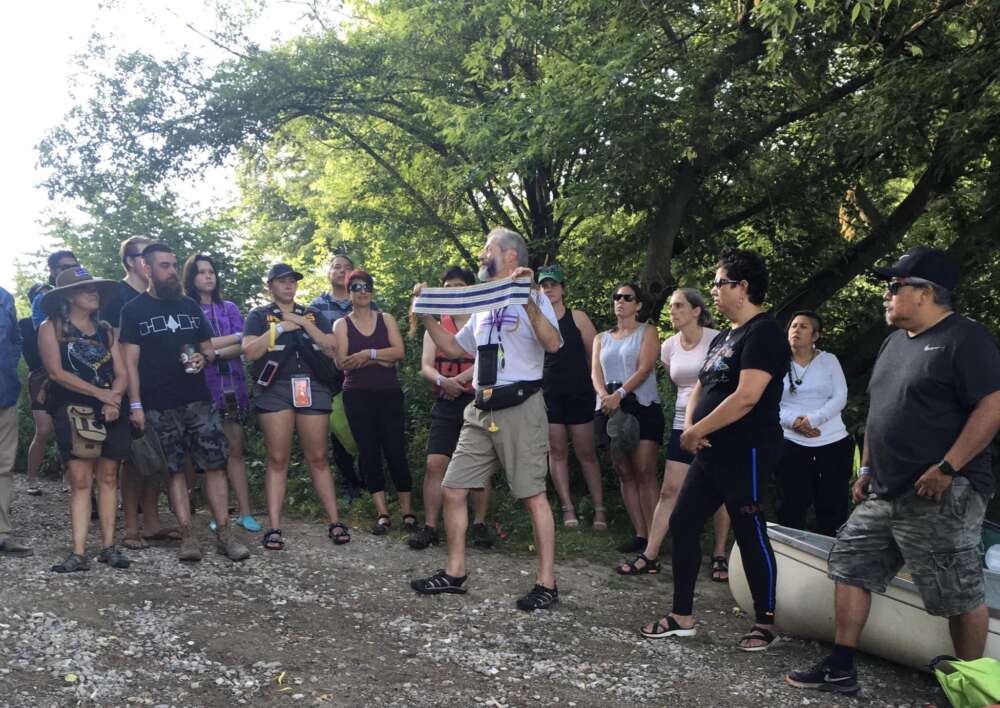 A group of fifteen gather outside around a demonstrator, who holds a beaded wampum belt. The belt has two rows of parallel purple lines against a white background.