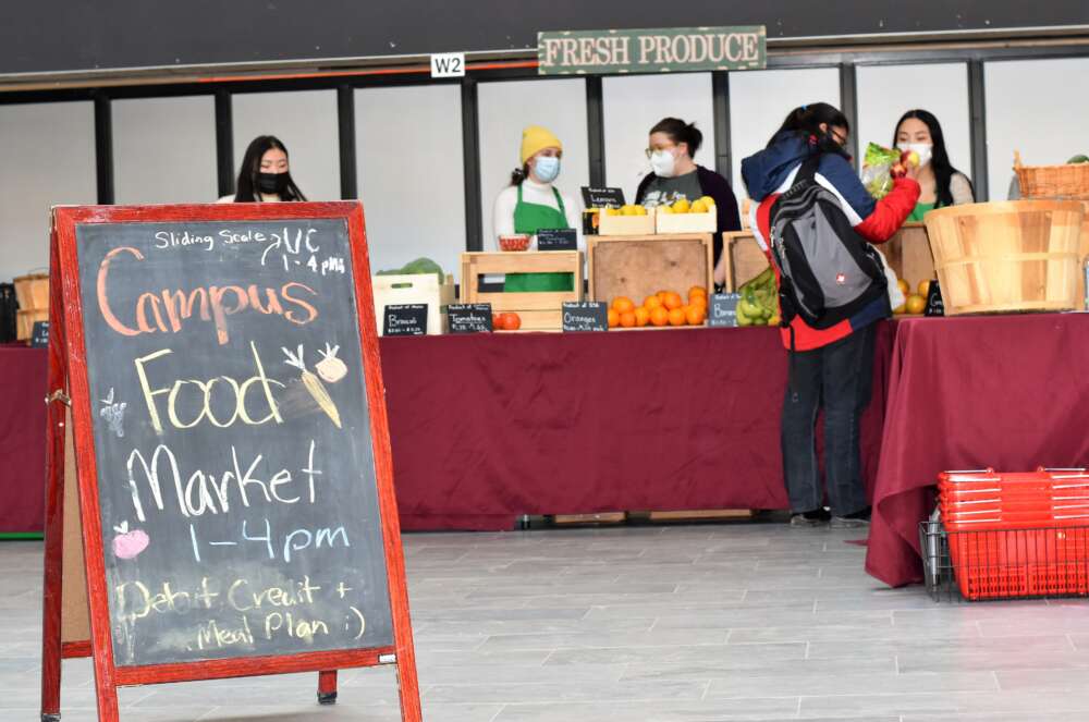 The campus food market, with a sign that reads "Campus Food Market, 1-4 pm" with a table of fresh produce and people speaking to one another in the background.