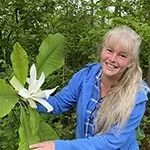 A woman holds up a white flower in a large green plant while out in the woods.