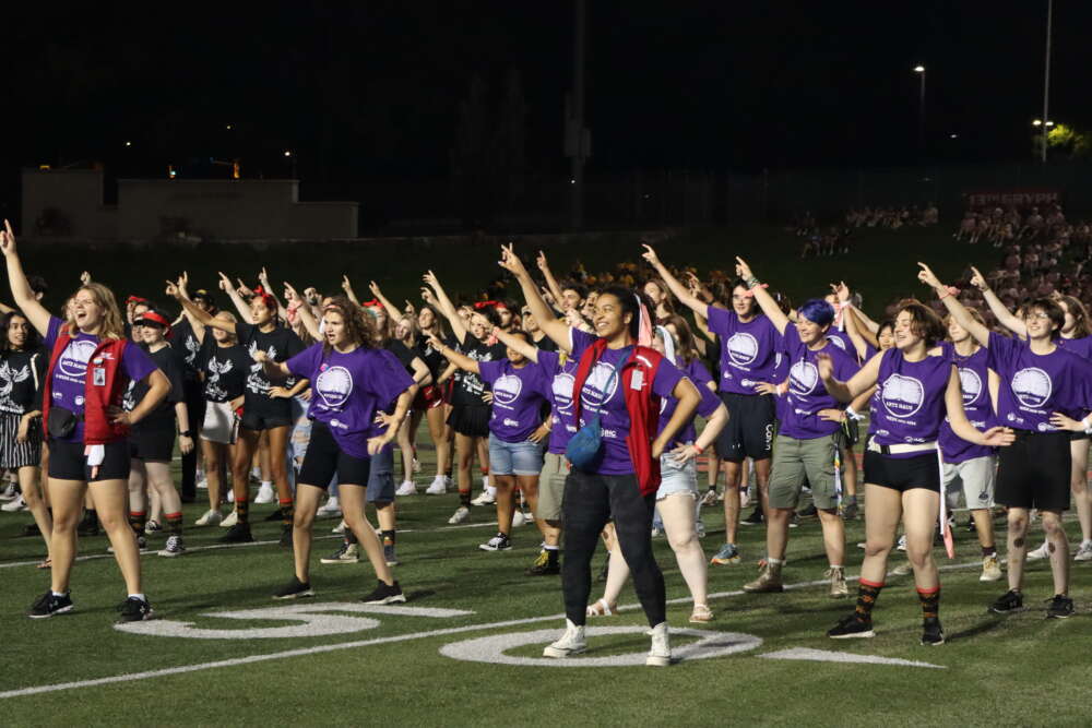 a team of students in dark purple shirts performs on a turf football field