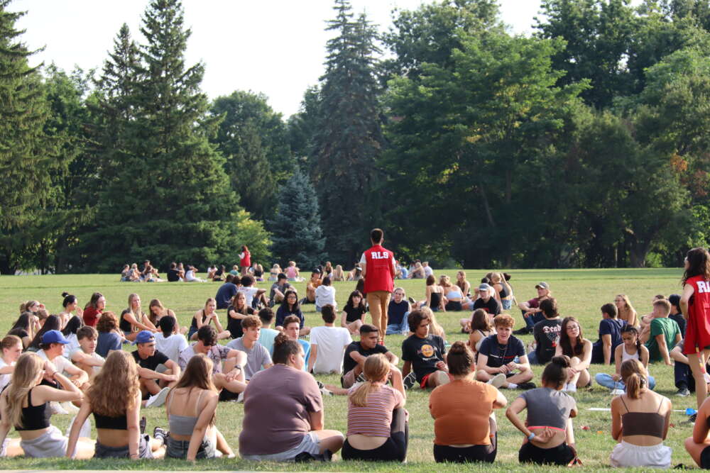 incoming university students sit in rows on the grass and receive information from standing volunteers