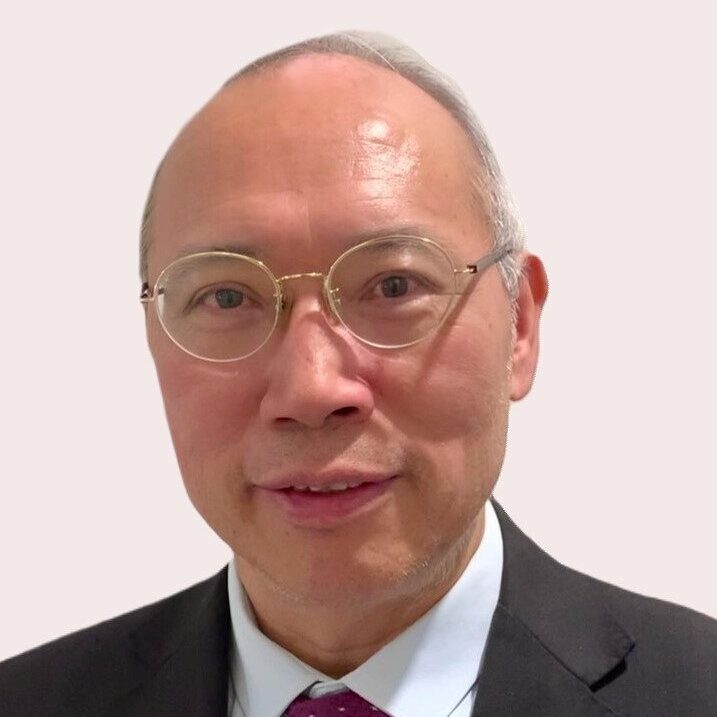 A person with wire-framed glasses wearing a suit and tie looks into the camera in front of white background.