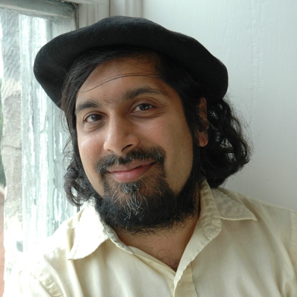 A man with long, dark curly hair and a beard wears a black beret and wears a cream-coloured collared shirt while smiling against the backdrop of a white wall and window.