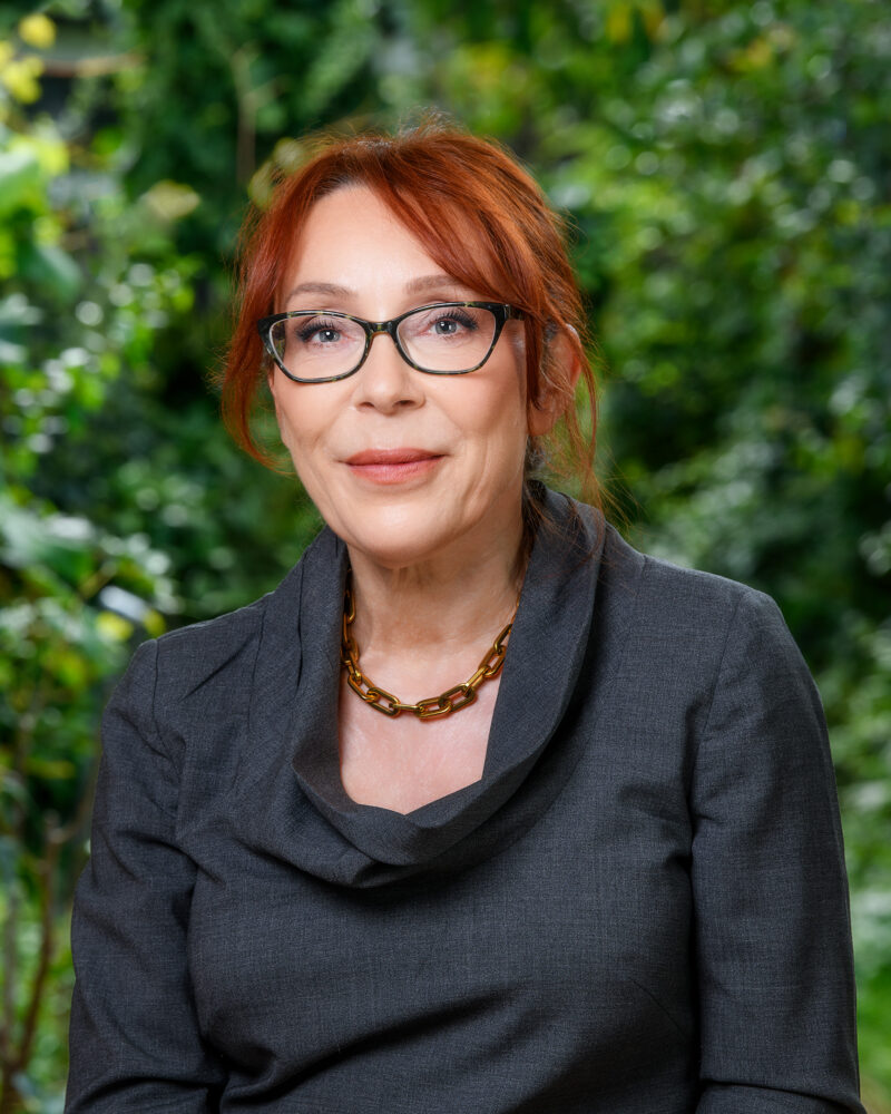 A person with red hair and black glasses wearing a charcoal dress and a gold chain necklace smiles into the camera in front of a background of greenery.