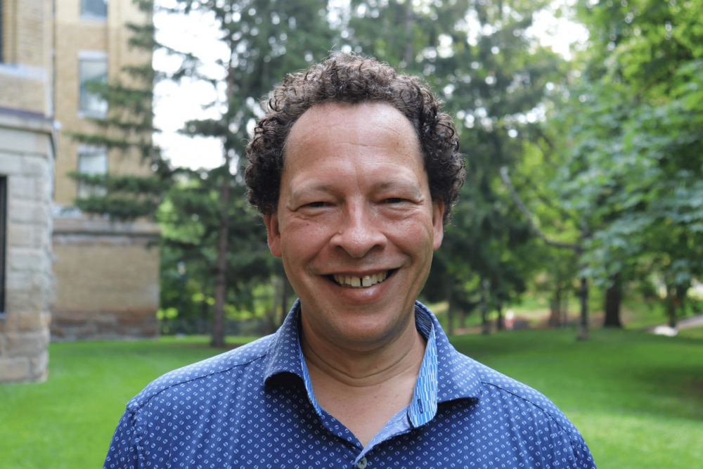 A person with short, dark curly hair wearing a collared shirt with blue pattern smiles into the camera standing on the U of G campus in front of a stone building and some trees.