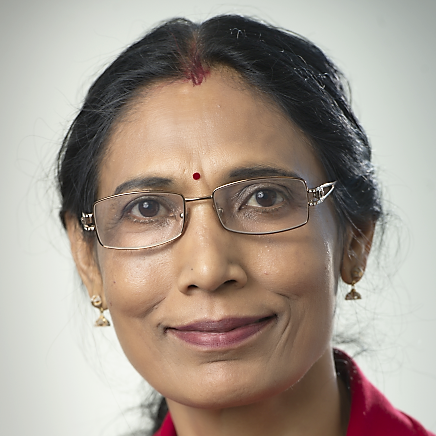 A person with black hair tied back and wire frame glasses wearing a red collared-shirt and coloured necklaces and earrings smiles into the camera against a white background.