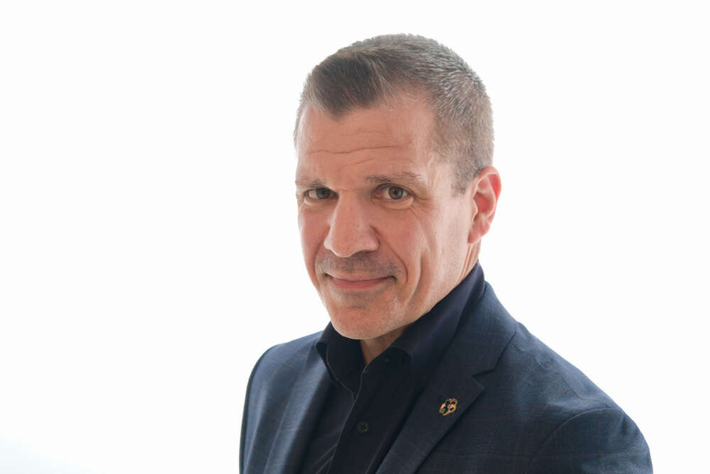 A man with short dark hair wearing a black collared shirt and suit jacket looks into the camera with a white background.