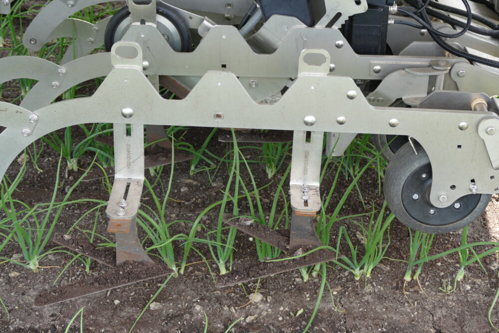 Close-up of the cultivator equipped on Farm Droid. Gray metal bars with ridges over them are parallel to the wheels underneath. Attached to the gray bars are blade-like cultivators.