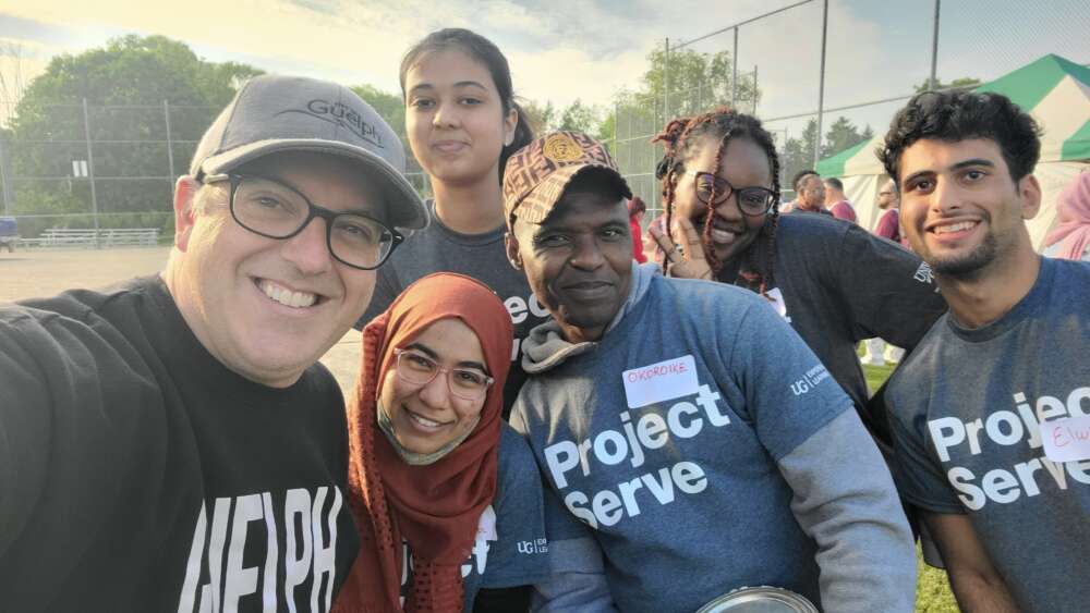 a group poses for a selfie, wearing grey shirts reading project serve with a baseball diamond in the background