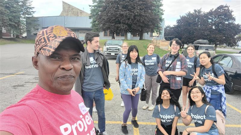 a Black man in a pink shirt reading Project Serve and a baseball cap takes a selfie with a group of students, all wearing grey shirts reading Project Serve