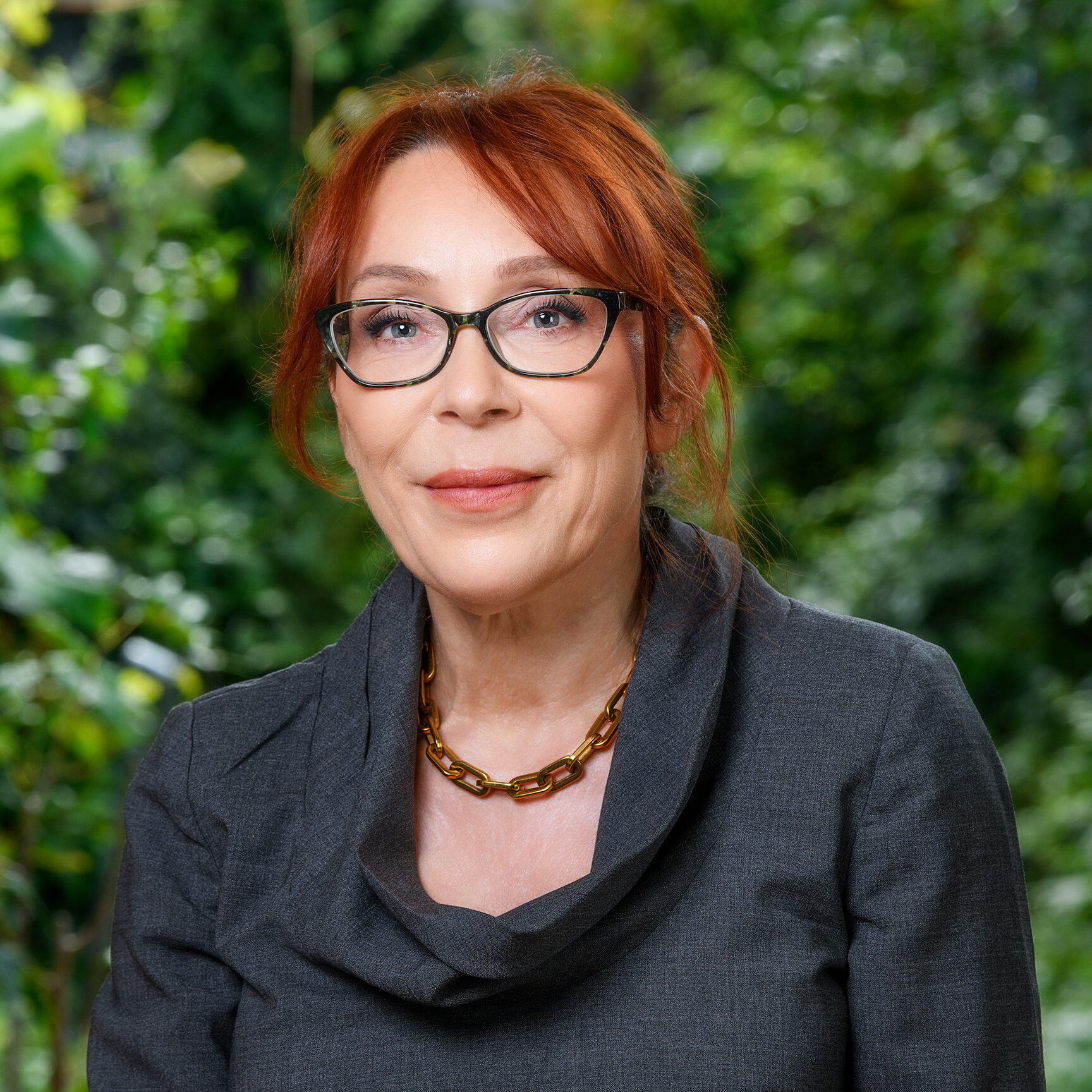 A person with red hair and black glasses wearing a charcoal dress and a gold chain necklace smiles into the camera in front of a background of greenery.