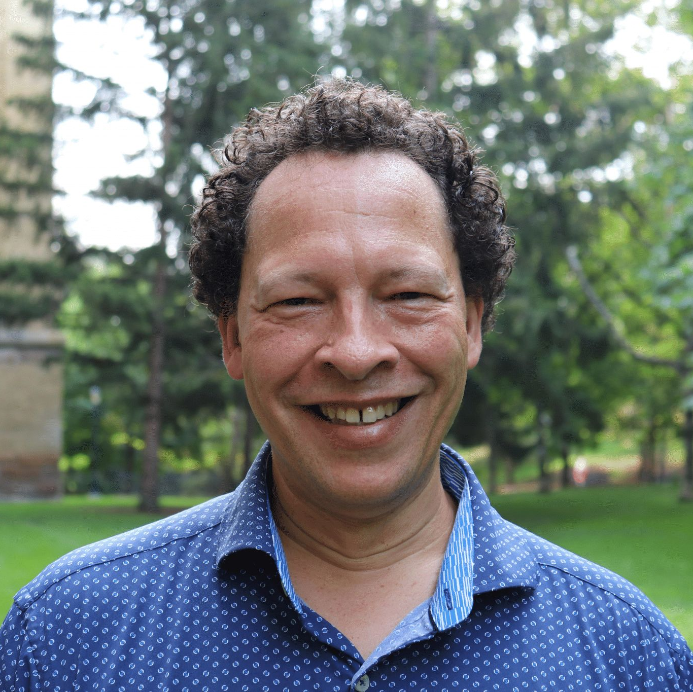 A person with short, dark curly hair wearing a collared shirt with blue pattern smiles into the camera standing on the U of G campus in front of a stone building and some trees.