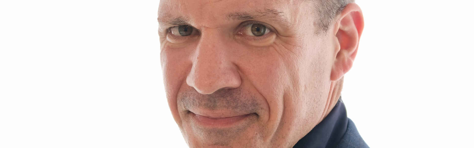 A man with short dark hair wearing a black collared shirt and suit jacket looks into the camera with a white background.