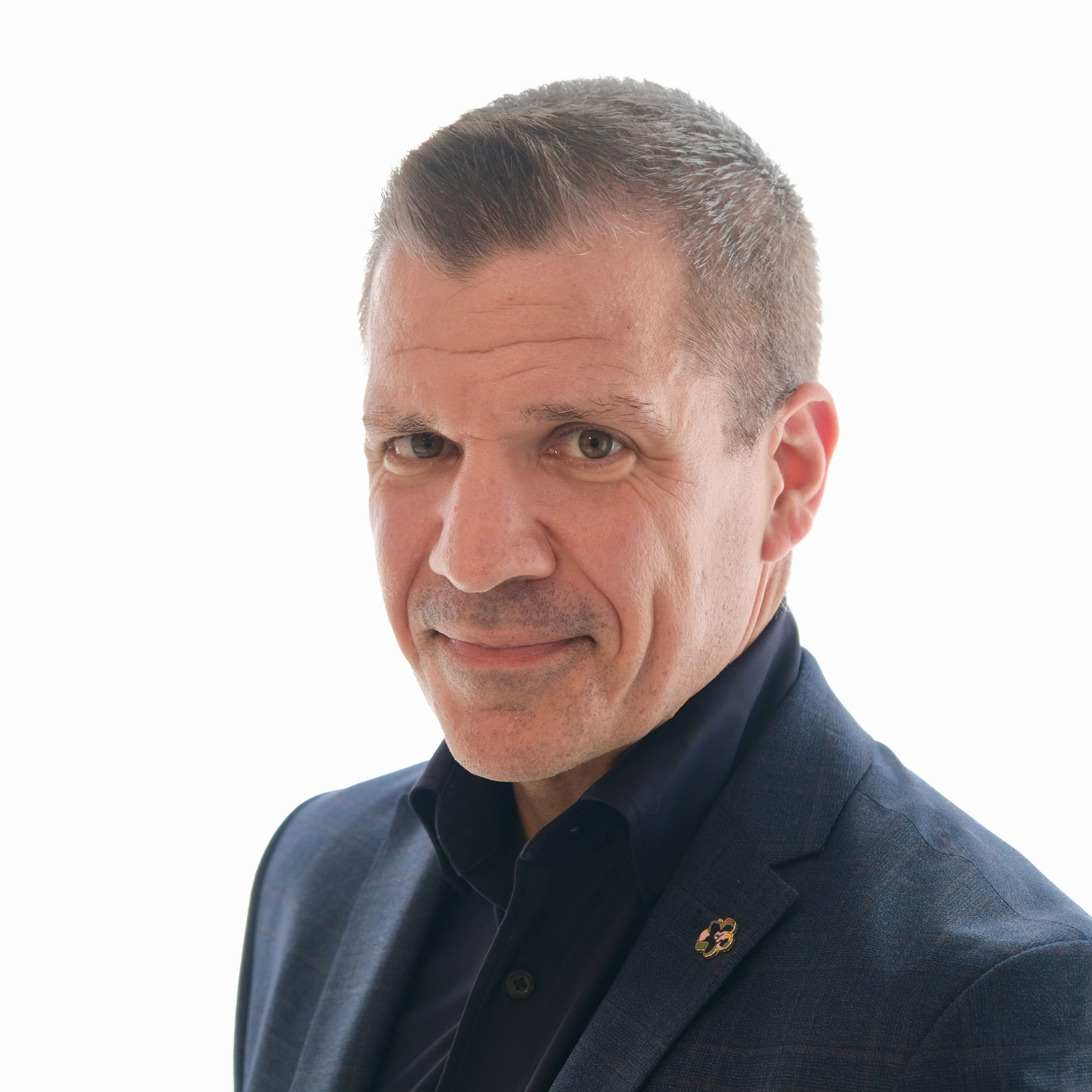 A man with short dark hair wearing a black collared shirt and suit jacket looks into the camera with a white background.