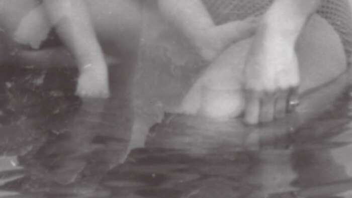 A blurry black and white photo with a person dipping a water pitcher into water, beside the feet of a small child.