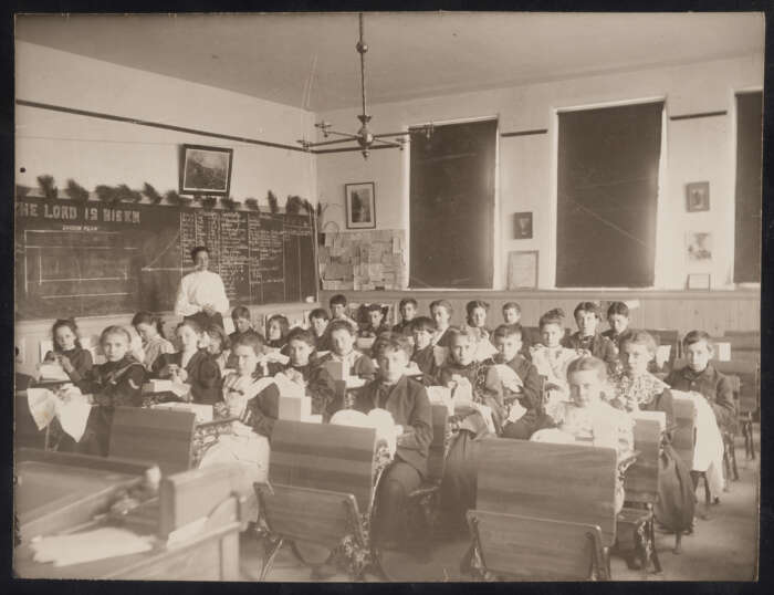 People sit in an old schoolroom in a black and white photo.