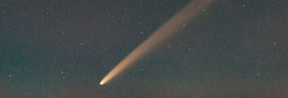 a comet streaks from the top right to bottom left corner of the night sky, bright against a dark background of stars
