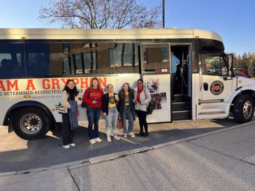 People in U of G clothing stand outside a white U of G bus in a parking lot.