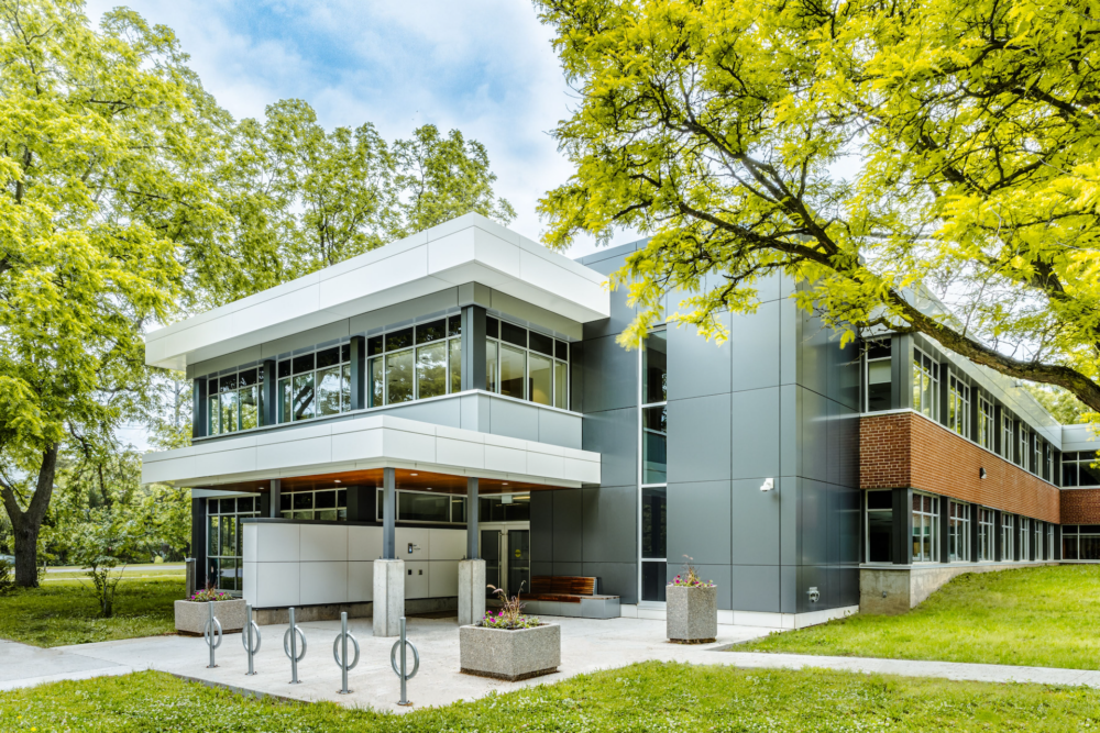 A two-story building surrounded by green trees, with a mix of brick and gray cladding on its facade. The design is modern, featuring large windows and clean, angular lines. The entrance area is sheltered by a white overhang supported by sleek columns, with a bike rack and planters placed outside. The building is surrounded by grass and trees.