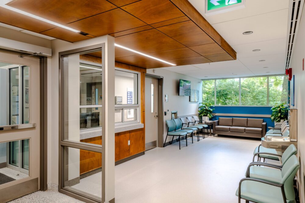 A modern waiting room with wood-paneled ceilings and bright lighting. The space features rows of light blue and beige chairs lining the walls, with a brown leather sofa positioned at the far end beneath a large window. There are two water fountains on the right side, and the walls are painted in a combination of beige and blue. The room has a few indoor plants and a flat-screen TV mounted on the wall. The window offers a view of lush green trees outside.