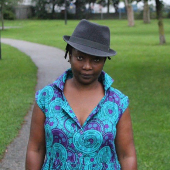 headshot of marsha hinds myrie, walking down an outdoor path wearing a fedora and bright blue and purple shirt.