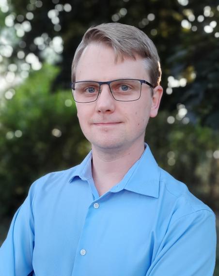 a man with glasses wearing a light blue shirt poses for a headshot in front of a green background