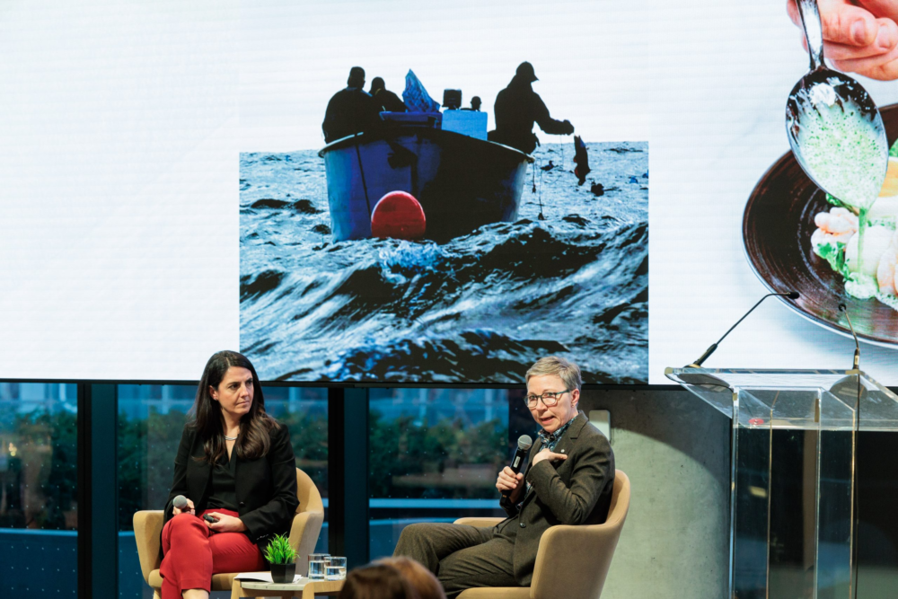 Two people on stage at a panel discussion, one holds a mic and addresses an audience. A projector screen with a photo of a boat and water and an image of food is behind them.