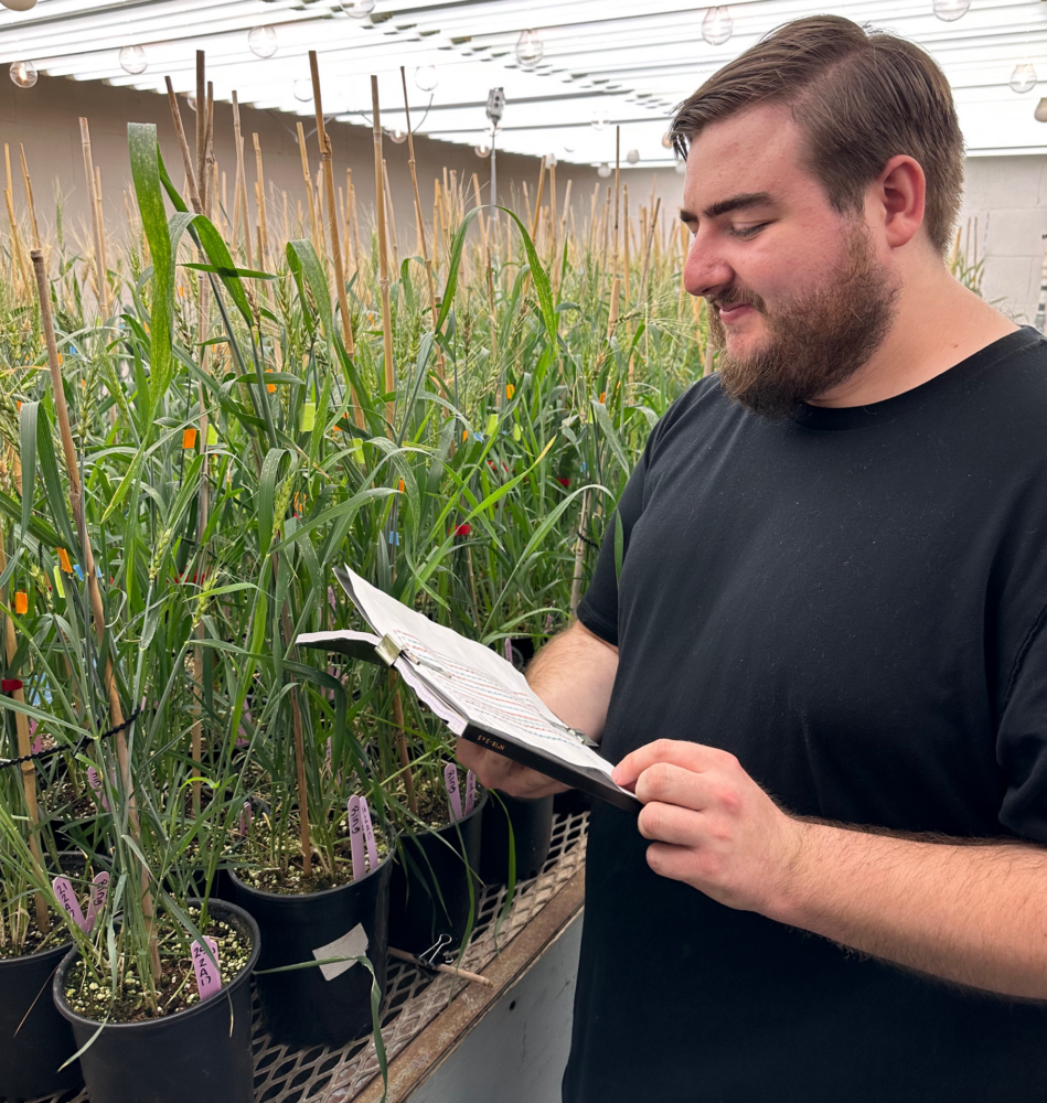 Connor Belot holding a notebook, smiling, beside rows of green plants growing in a well-lit lab