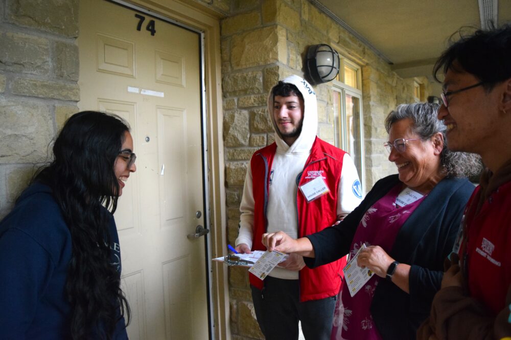 a young woman in dark blue stands beside a door, facing three people in red vests, a woman in glasses hands her a pamphlet