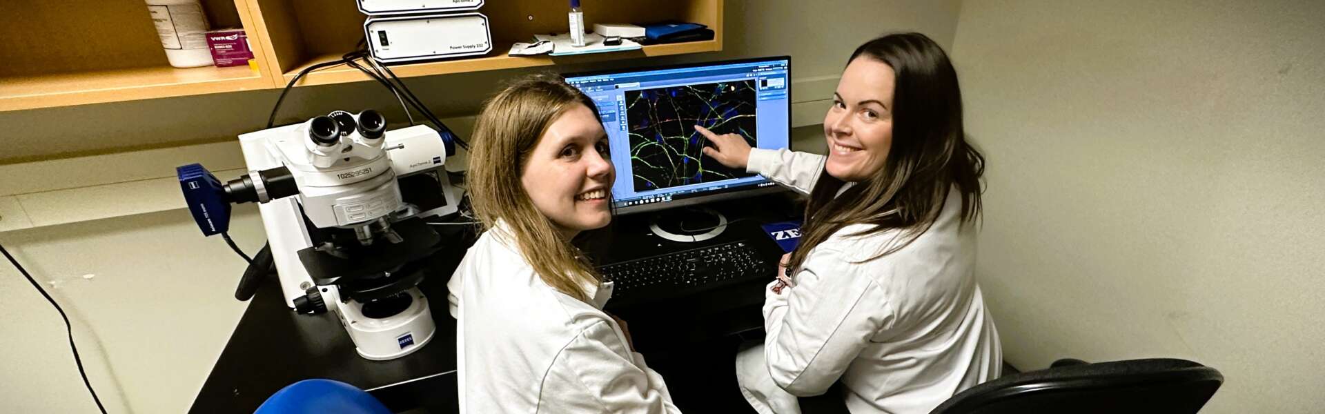 two women in lab coats sit in desk chairs in front of a computer screen, which shows samples from neurons