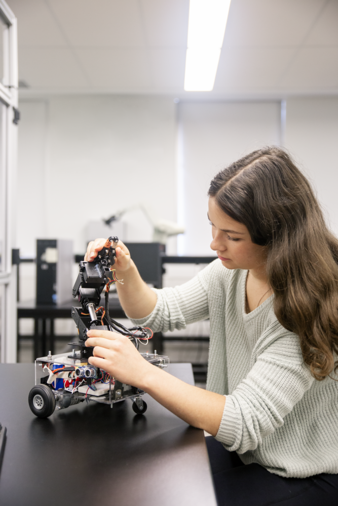 A student wearing a light-colored sweater is sitting at a table in a lab. She is assembling and adjusting a robotic arm attached to a wheeled robotic platform. The workspace is filled with electronics, wires, and equipment, emphasizing an engineering or robotics environment.