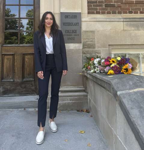 A person with long brown hair wearing a navy pant suit and white blouse and shoes stands on the steps of the Ontario Veterinary College with the brick building and wood and glass doors behind them.