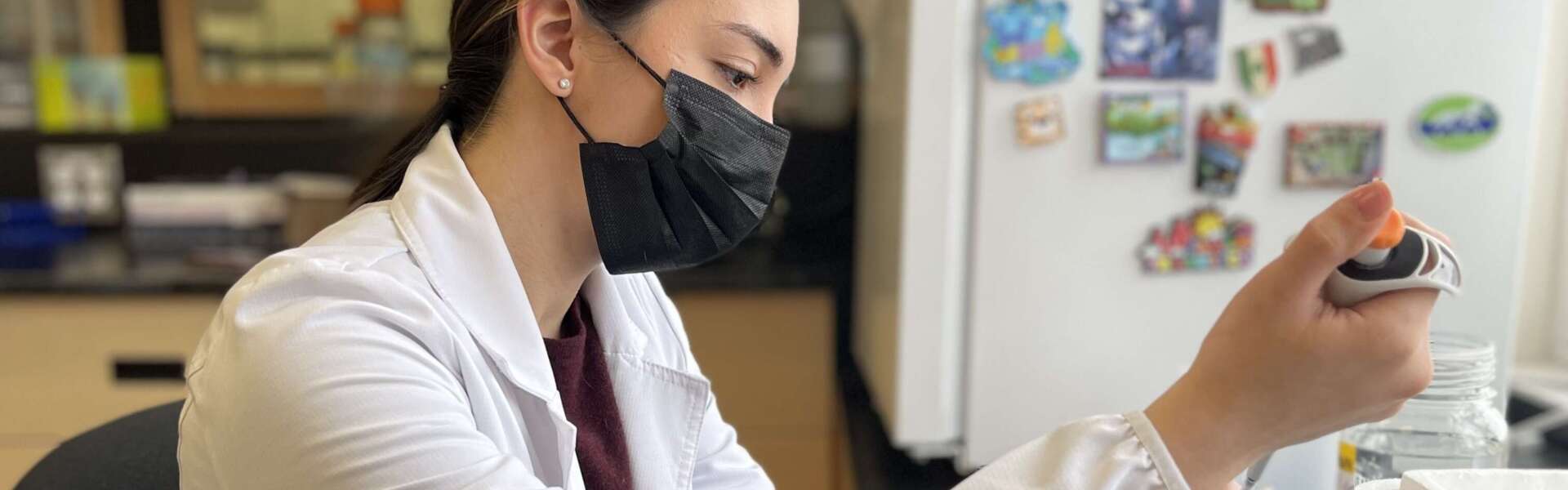 A person with brown hair wearing a black face mask and a white lab coat sits at a desk and transfers specimen.