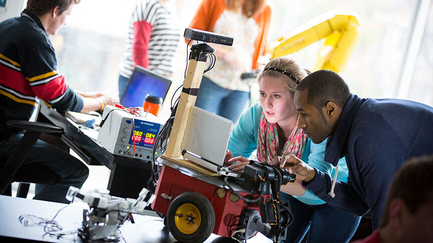 Two people work collaboratively on a project involving a small wheeled robot. The robot is equipped with sensors and a mounted camera on top. The duo is focused on a laptop screen connected to the robot. In the background, another person works at a desk with technical equipment.