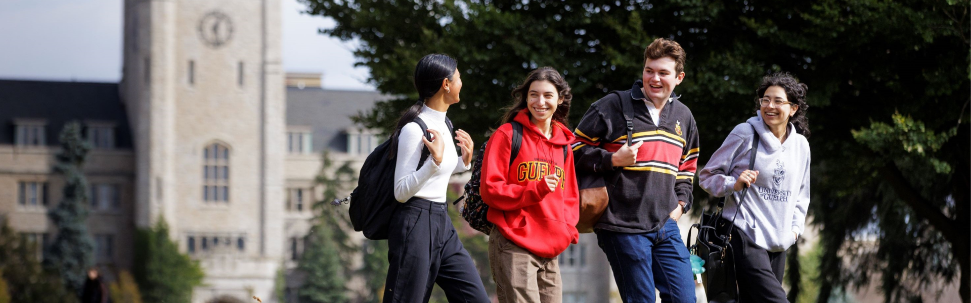 Four students walk in a line smiling at each other, wearing University of Guelph sweaters, against a campus background.