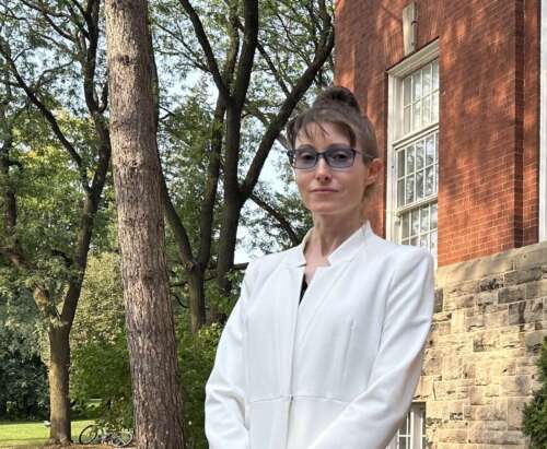 A person in a white overcoat, brown hair and glasses stands in front of a red brick building with trees surrounding.
