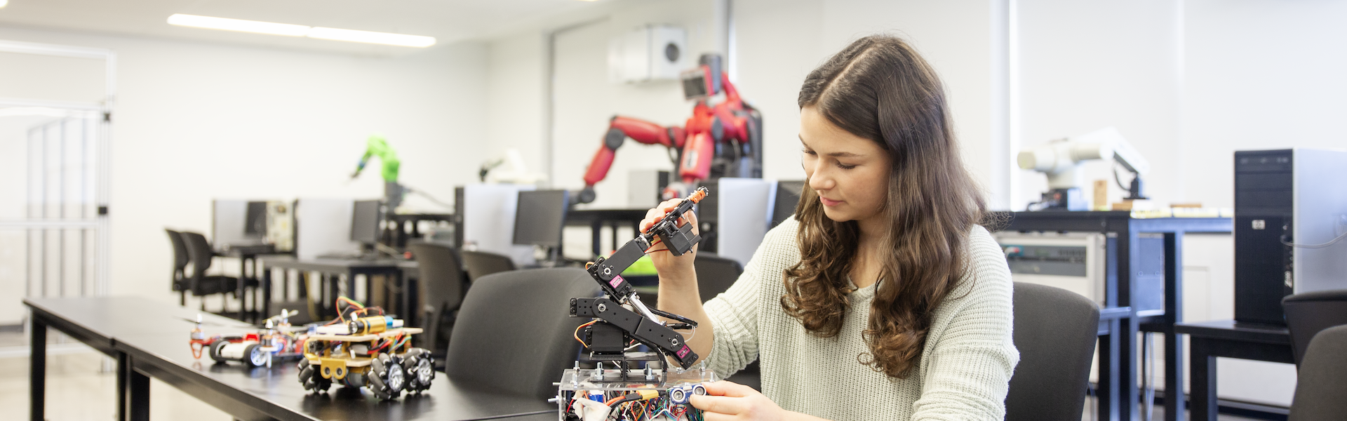 A student is seated in a lab surrounded by computers and robotic equipment. She is working on a wheeled robot with a robotic arm. Other robots and machinery are visible in the background, creating a modern robotics lab atmosphere.