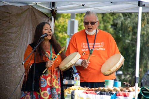 Elder Dan Smoke and Mary Lou Smoke drum outdoors at an event.