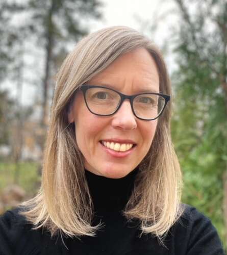 A person with shoulder-length blonde and brown hair wearing black frame glasses and a black turtleneck smiles into the camera standing in front of a background of tall green trees.