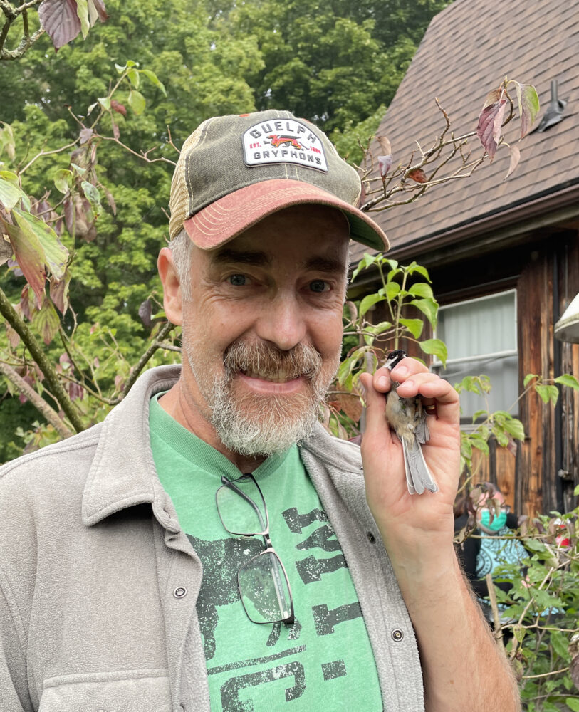 a man in a green shirt and grey jacket wears a grey and red ballcap, he's holding a small grey bird in his left hand