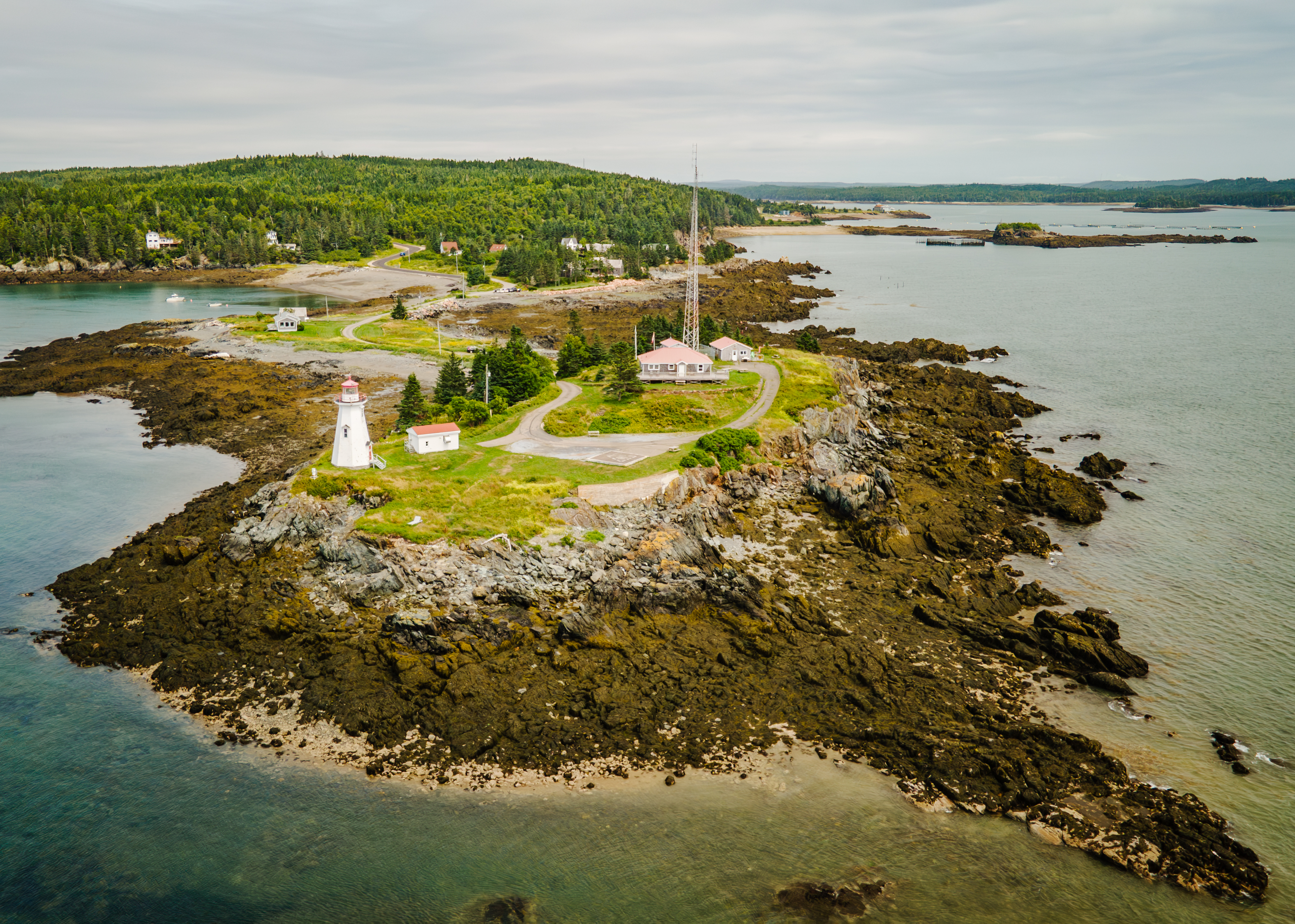 An aerial image of a scenic coastal area with a lighthouse prominently featured on a rocky peninsula. The landscape is rugged, with rocky shores extending into the water on either side. The lighthouse, painted white with red accents, stands near a few small buildings and a winding road leading towards a larger structure and a tall communication tower. The surrounding waters are calm, with a forested background stretching into the horizon.