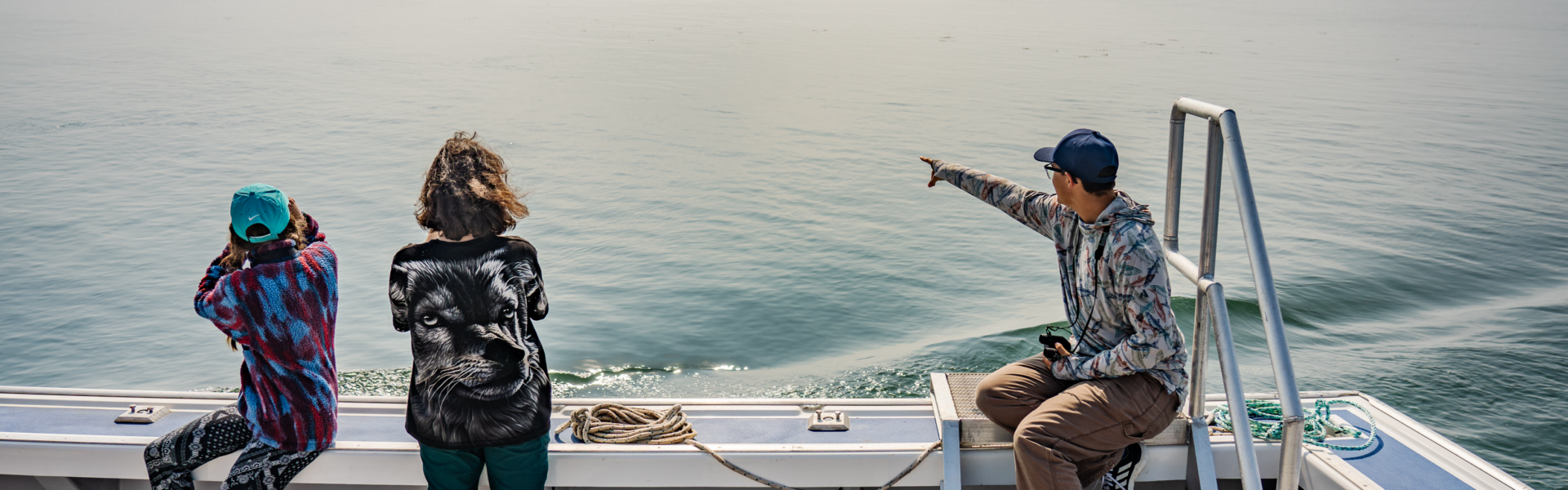 Three students on a boat, with two observing the water and one pointing towards the horizon. The scene is misty, with a calm sea stretching out in front of them.