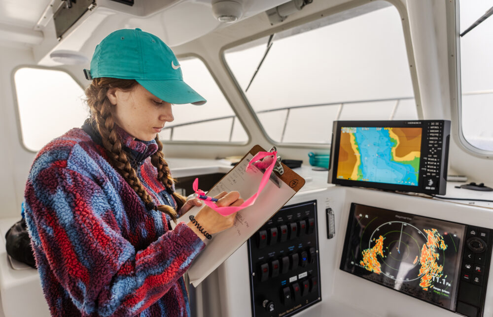 student wearing a turquoise cap and holding a clipboard, standing inside a boat’s control room. Marine navigation screens display maps and radar in the background.