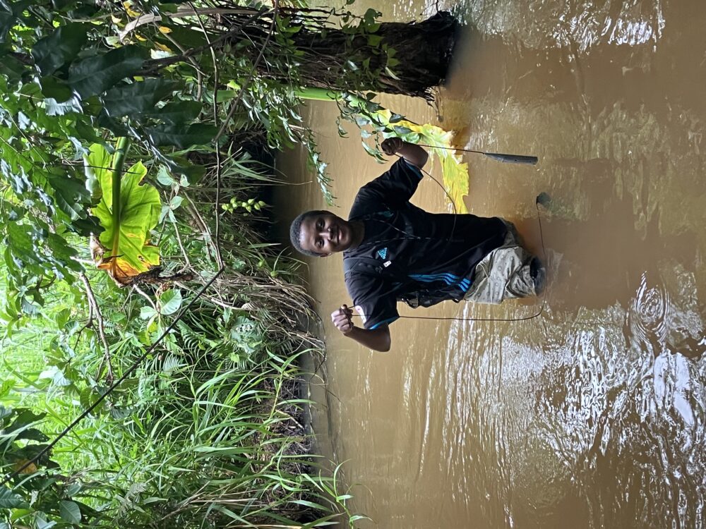 Amilcar Bisekere standing knee-deep in a muddy river, surrounded by lush greenery, wearing a black shirt and light shorts, holding sampling equipment while smiling at the camera