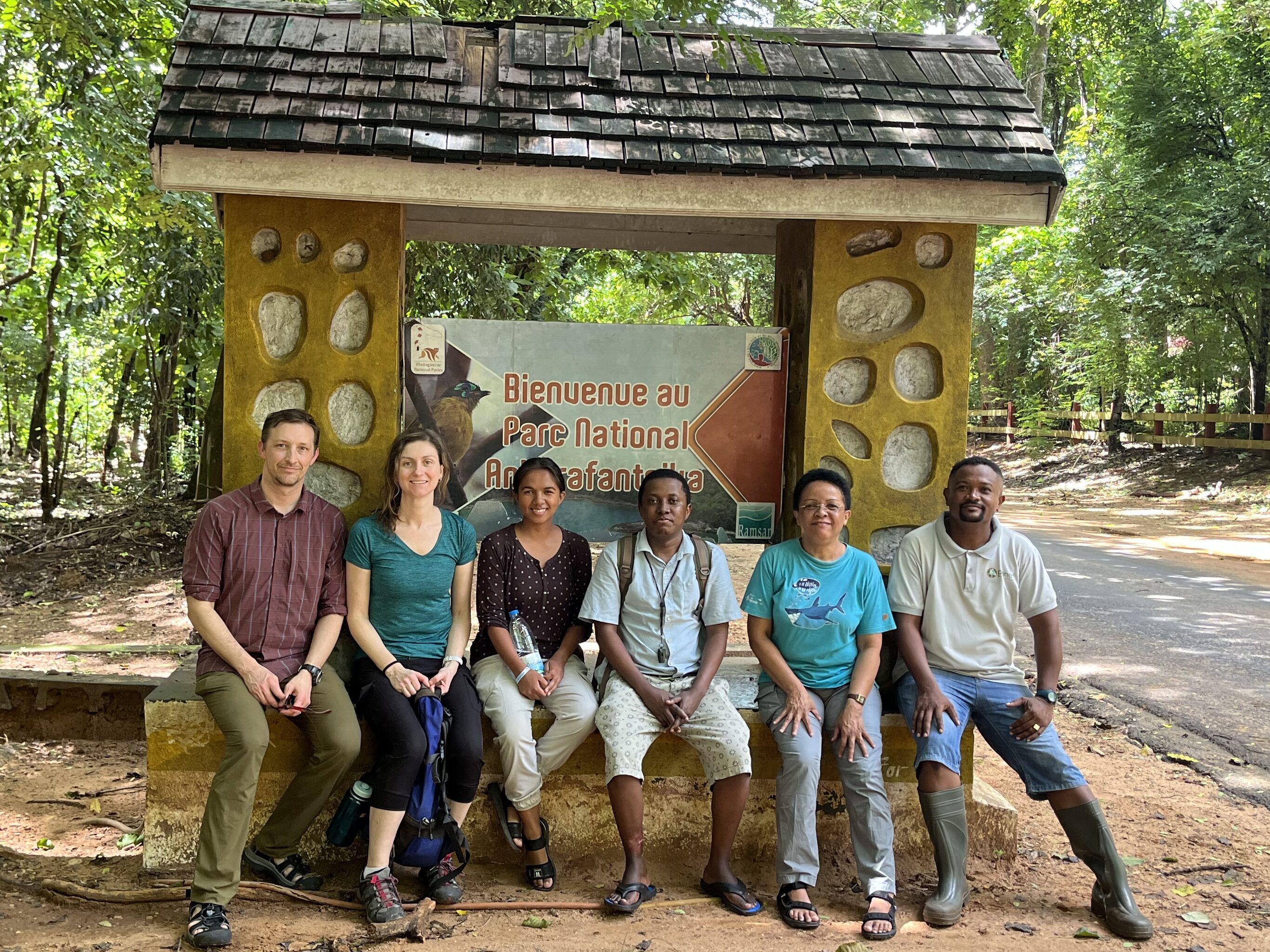 A group of individuals standing in an outdoor setting, seemingly just arriving at a natural reserve. The background features dense vegetation and a dirt path. Some members are holding bags and wearing casual, outdoor attire, indicating readiness for fieldwork or exploration. The scene captures the arrival of a research team at Ankarafatsika National Park after a long journey