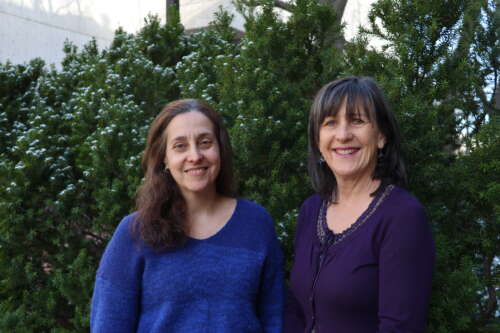 A person with long brown hair wearing a blue and white sweater stands smiling beside a person with shoulder length dark hair wearing purple in front of a green bush lightly dusted with snow.